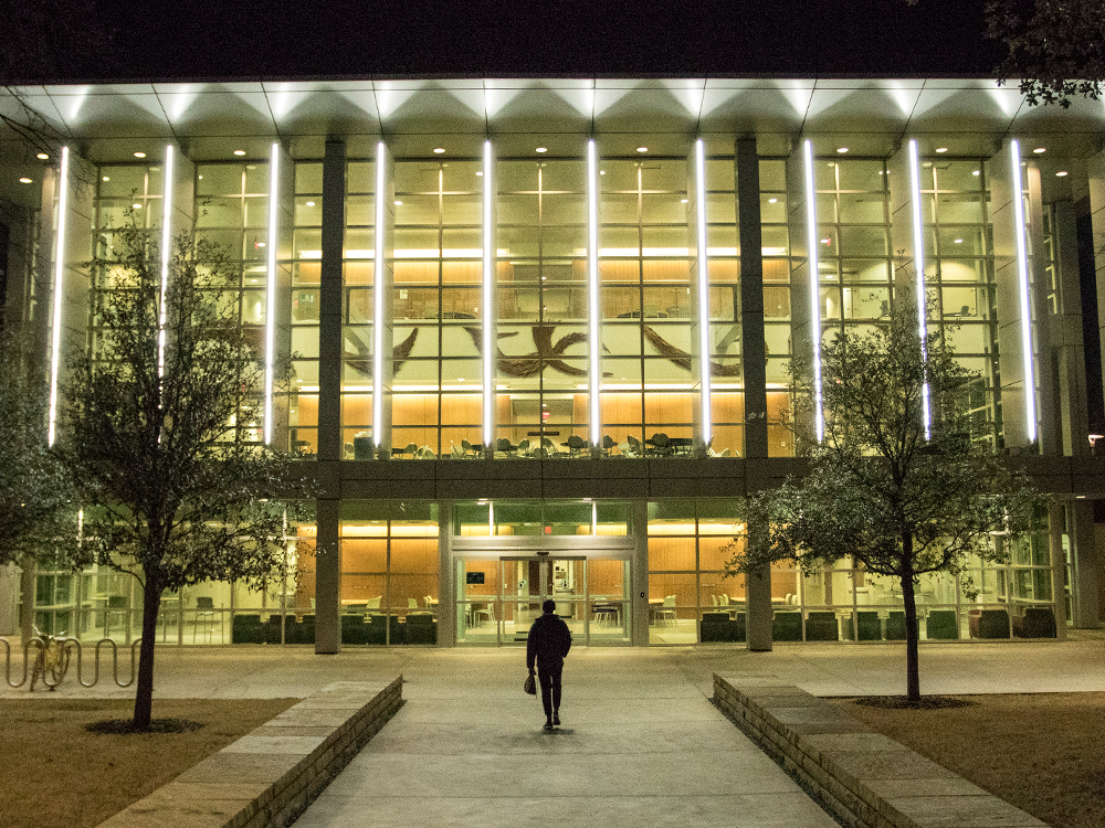 A lone student walks toward a lit-up Founders Building at night