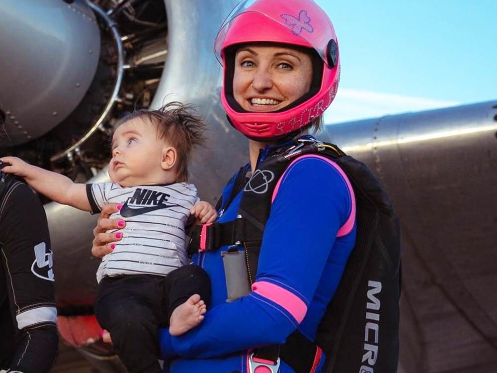 A woman in skydiving gear, a pink helmet and blue jumpsuit with pink piping, holds her son next two a sitting airplane.