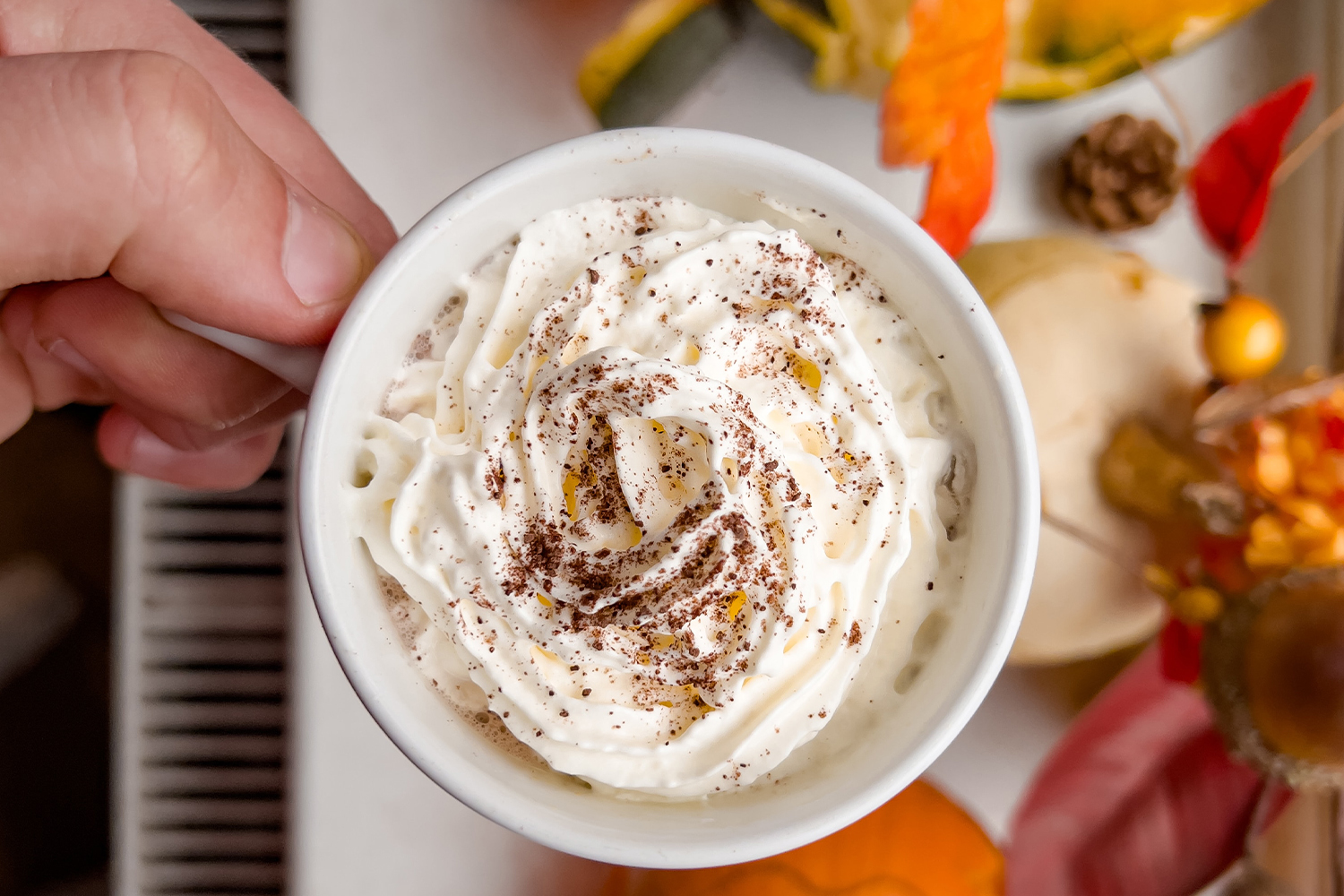 A close-up image of a person’s hand holding a pumpkin spice latte with fall decor below.